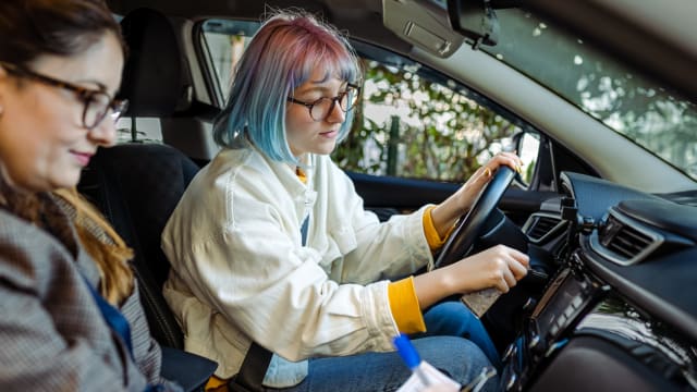 A young woman with colourful hair starting a car. A driving instructor is sitting next to her and taking notes.