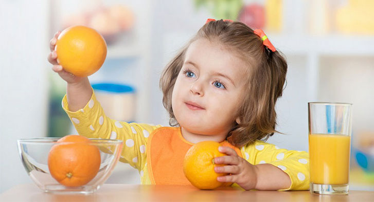 15 Motivos Para Tomar Suco de Laranja com Cenoura + Receitas Menina de 5 anos tomando suco de laranja e absorvendo vitamina C. Ela está com 2 laranjas na mão e um copo de suco de laranja ao lado.