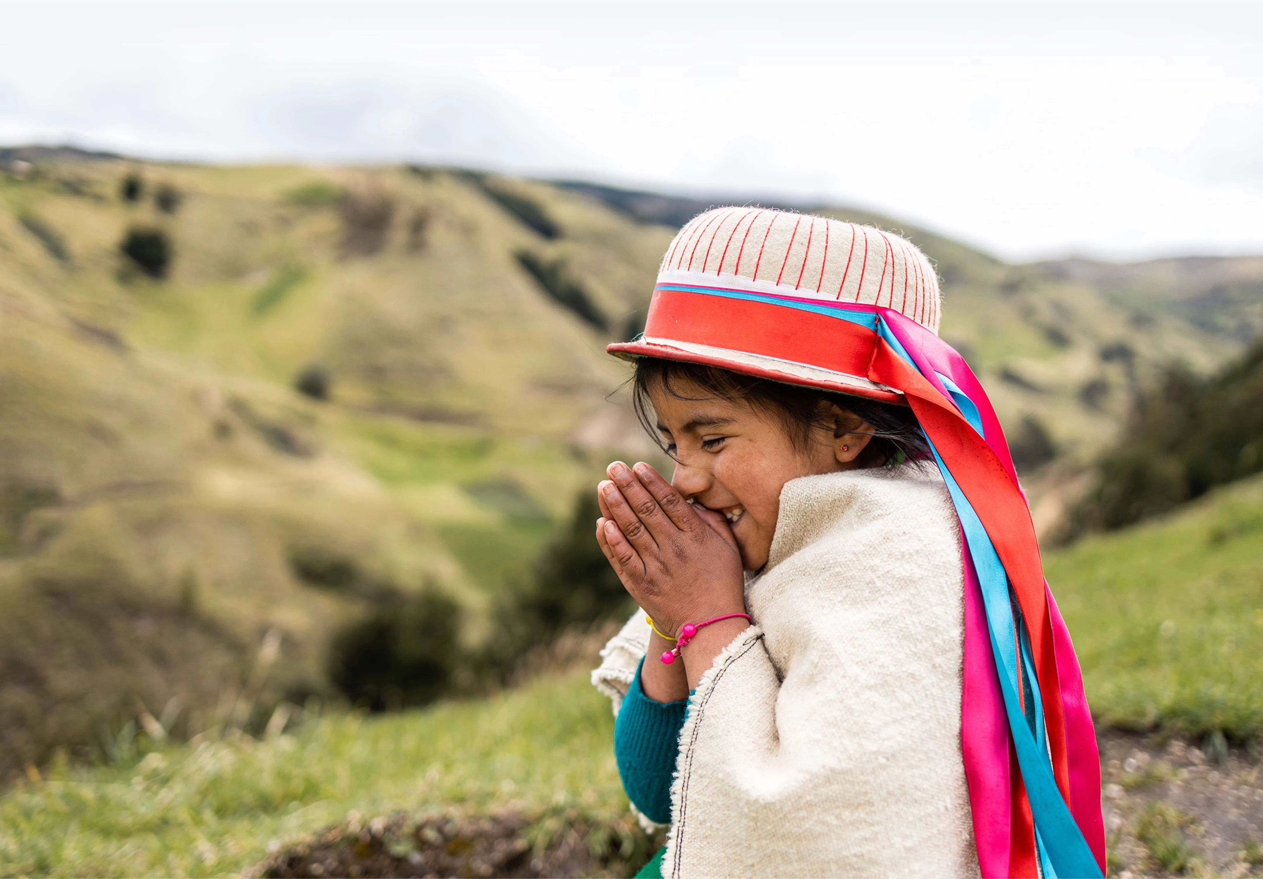 Image of girl in traditonal garb, praying with her hands to pressed together. She stands in a mountainous landscape