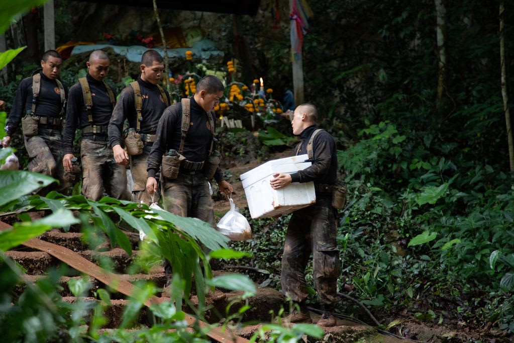 Four men in uniform walk down a path, the one in front holding a white box.