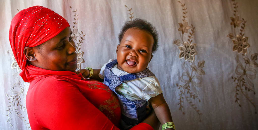 A Ugandan woman wearing a red shirt and head scarf smiles at the baby she holds in her arms.