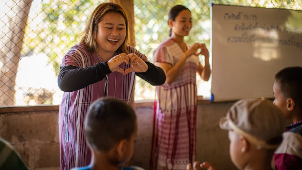 A woman smiles at a group of children and holds her hands in a heart shape.