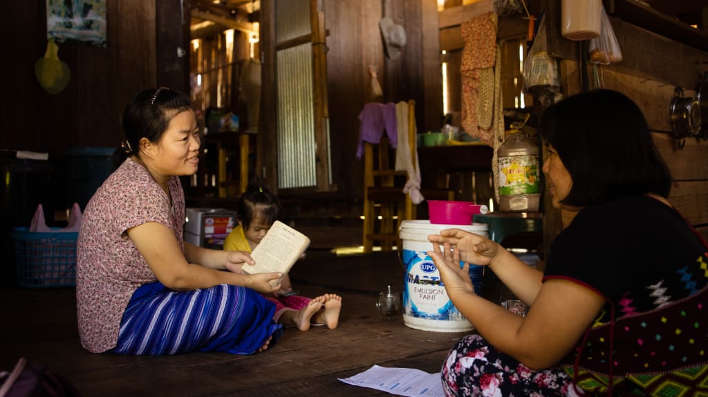 A woman in a patterned top and blue skirt skits on the floor with a book in her hands and talks to another woman.