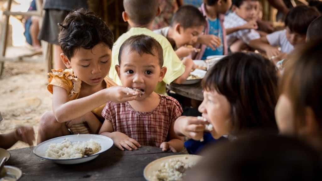 Children are sitting at wood tables eating rice. One child is feeding another child with their hand.