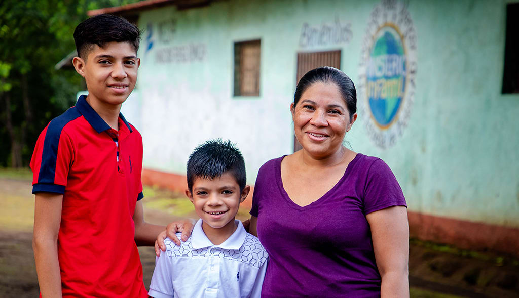 Holman and Joseph with their mother, Francis. She is wearing a purple t-shirt and they are standing in front of the Compassion centre.