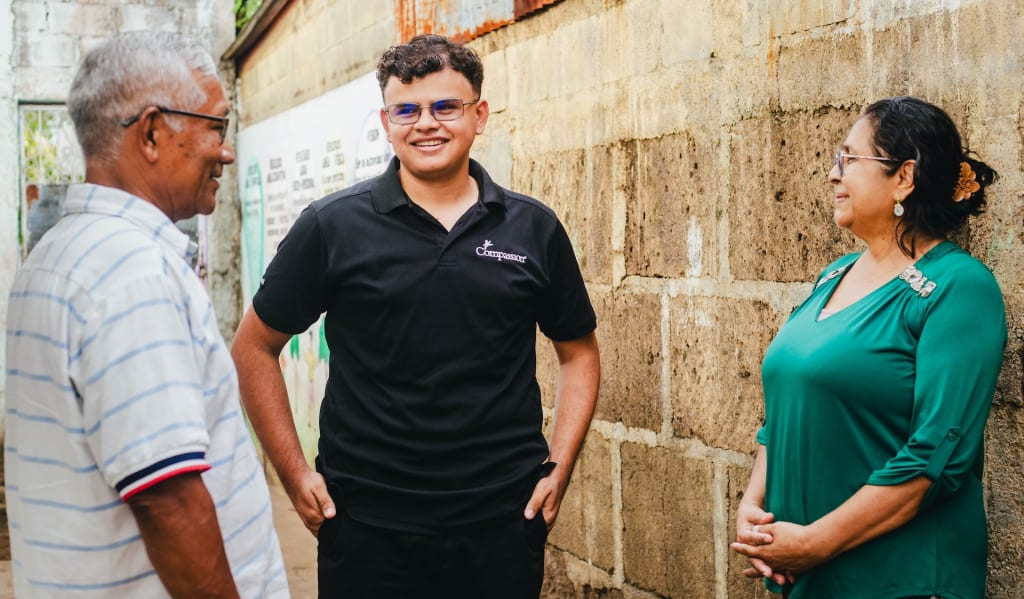 Three adults stand in front of a brick wall, smiling and looking at one another.