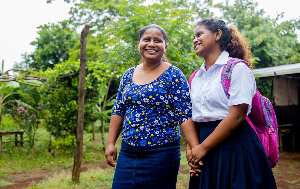 Crisli and her mom walking to school. Cristi wears a school uniform and a pink backpack. Her mother wears a blue skirt and a blue shirt.