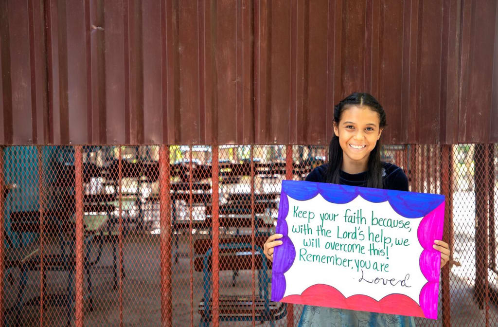 Little girl sits in front of a fence holding a sign that says, "Keep your faith because with the Lord's help, we will overcome this! Remember, you are loved."