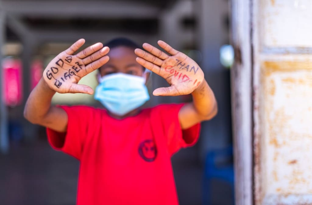 Little boy wearing a red shirt and a blue mask opens his hands to the camera. On his hands are written, "God is bigger than COVID-19."