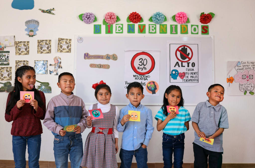 In Mexico, six children stand in a line at the front of a classroom. Each child is holding a drawing of a face representing a different emotion, and they are all acting out the emotions.
