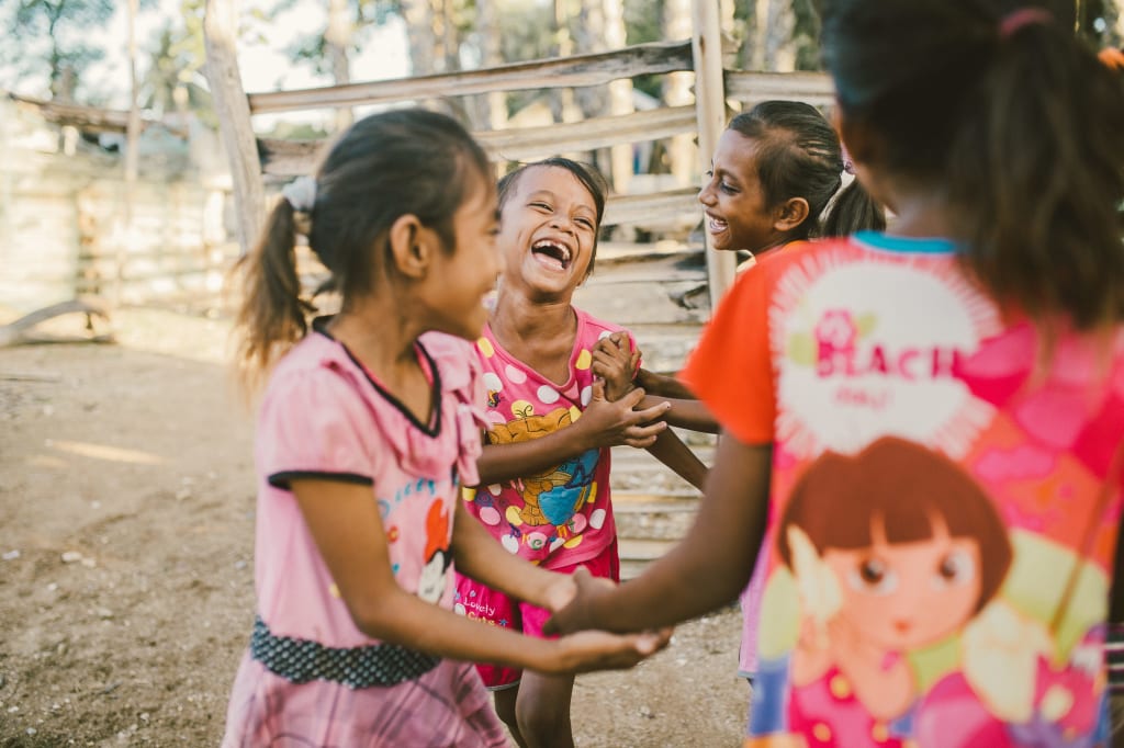 Talita, Julenda, Jani and Tersya, in pink and orange shirts, are outside laughing and playing. There is a fence behind them.