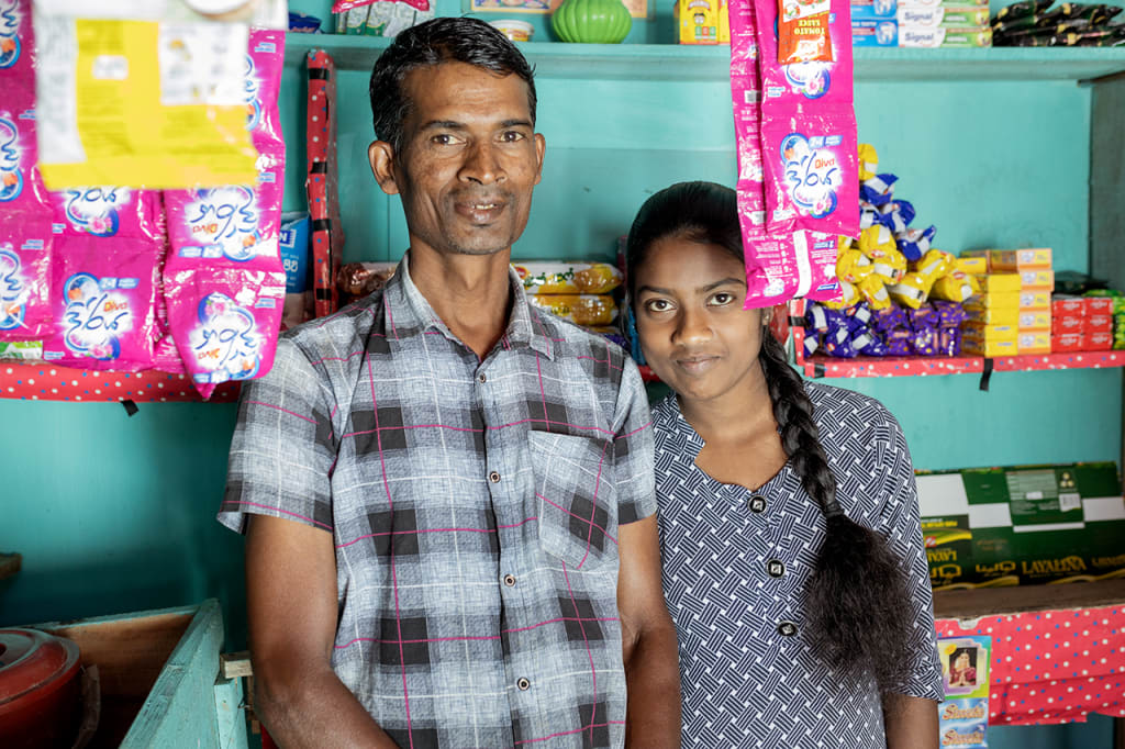 A man and a girl, his daughter, in a small shop. There is food behind them on shelves.