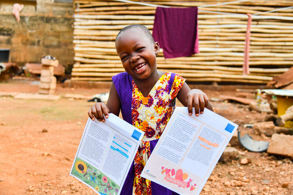 A girl in a colourful dress holds up two sheets of paper and laughs.