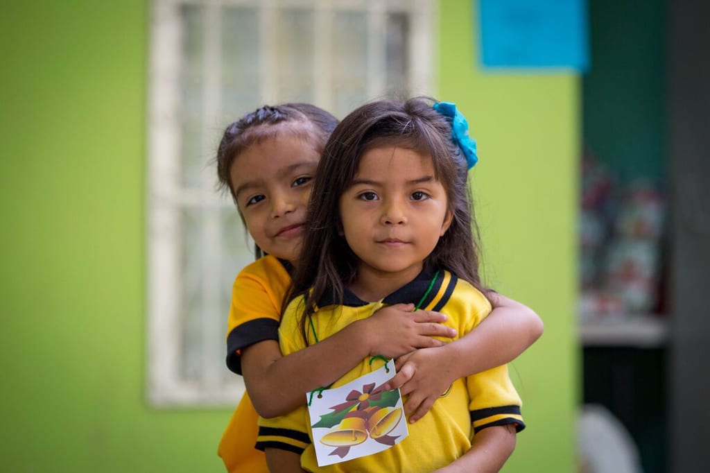 Two girls hug while looking at the camera. They are wearing yellow and standing in front of a green wall.