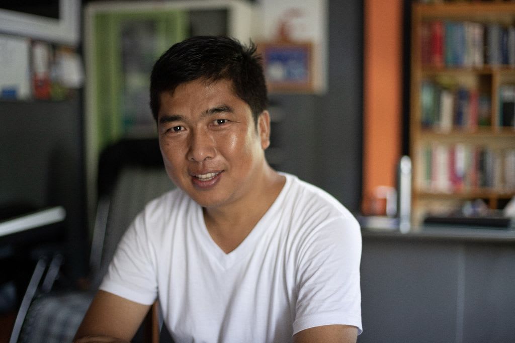 A man in a white shirt sits at a table and smiles at the camera.