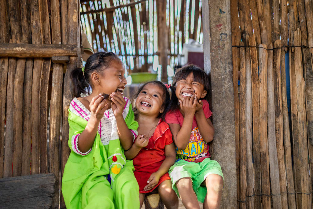 Eldimar is wearing a bright green dress. She is sitting in the doorway of her home with two of her sisters. They are all eating cake.