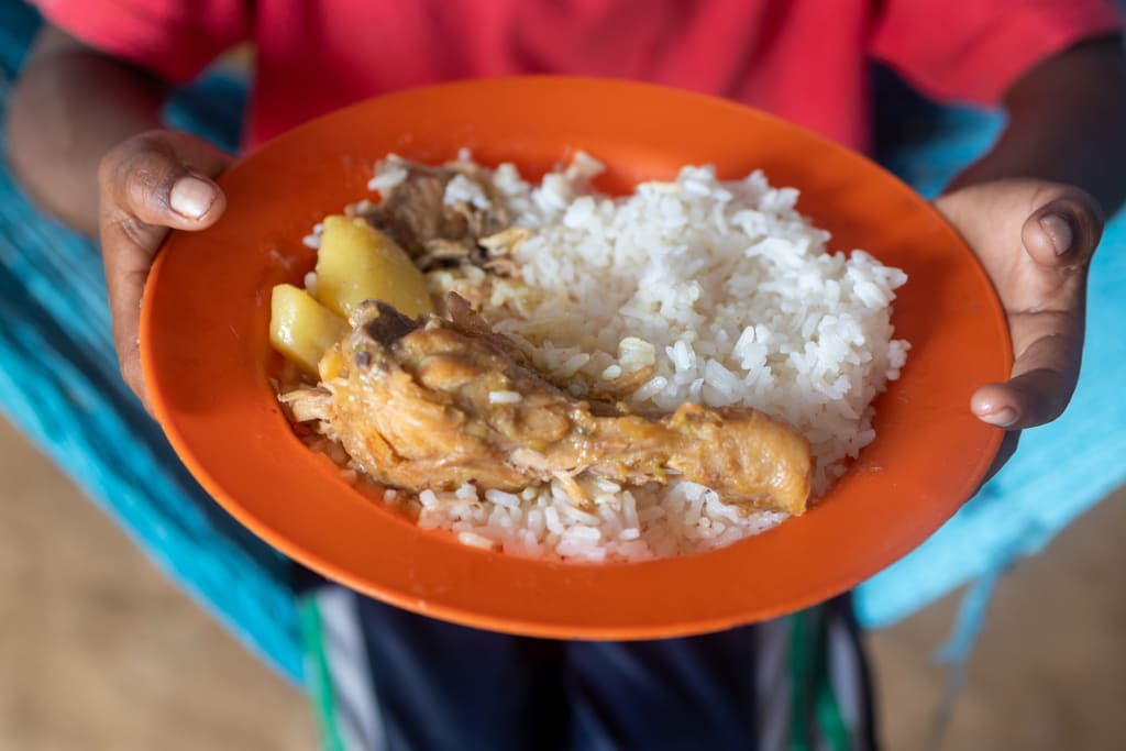 An orange plate is held by a child, on it is some cooked white rice, a small piece of chicken and a few vegetables in sauce.