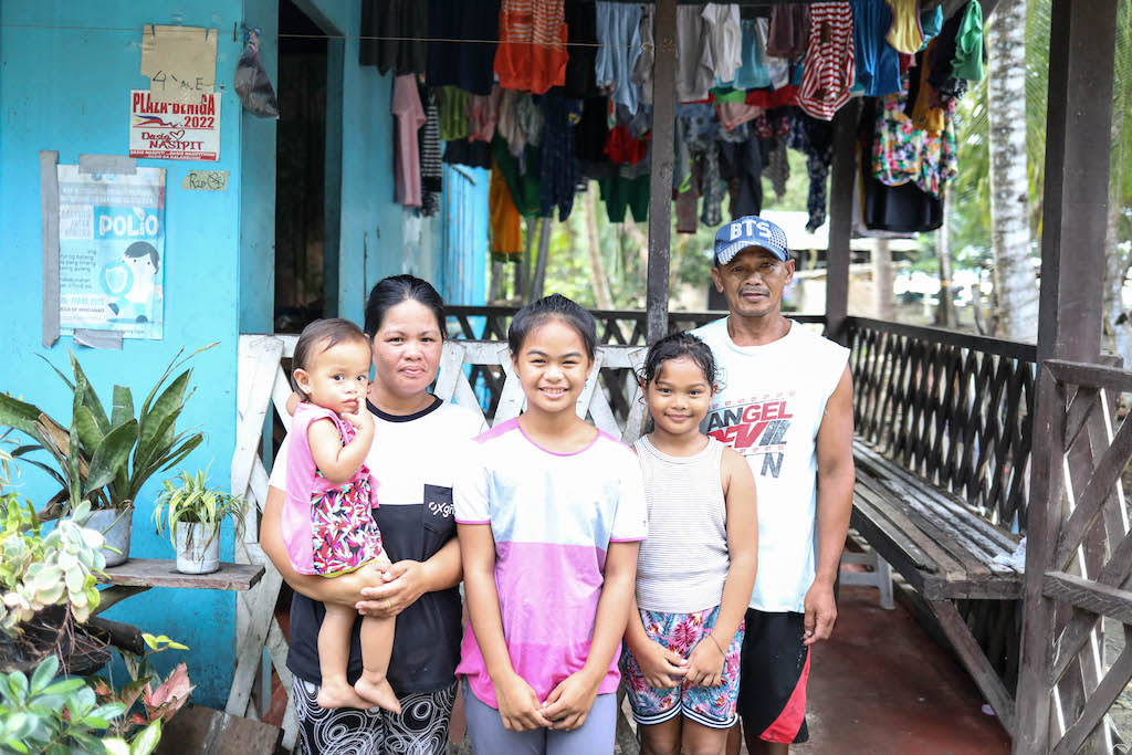Kareen is wearing a pink, white, and gray striped shirt. She is standing outside her home with her mother, family, and two siblings. There are clothes hanging on a clothesline above them.