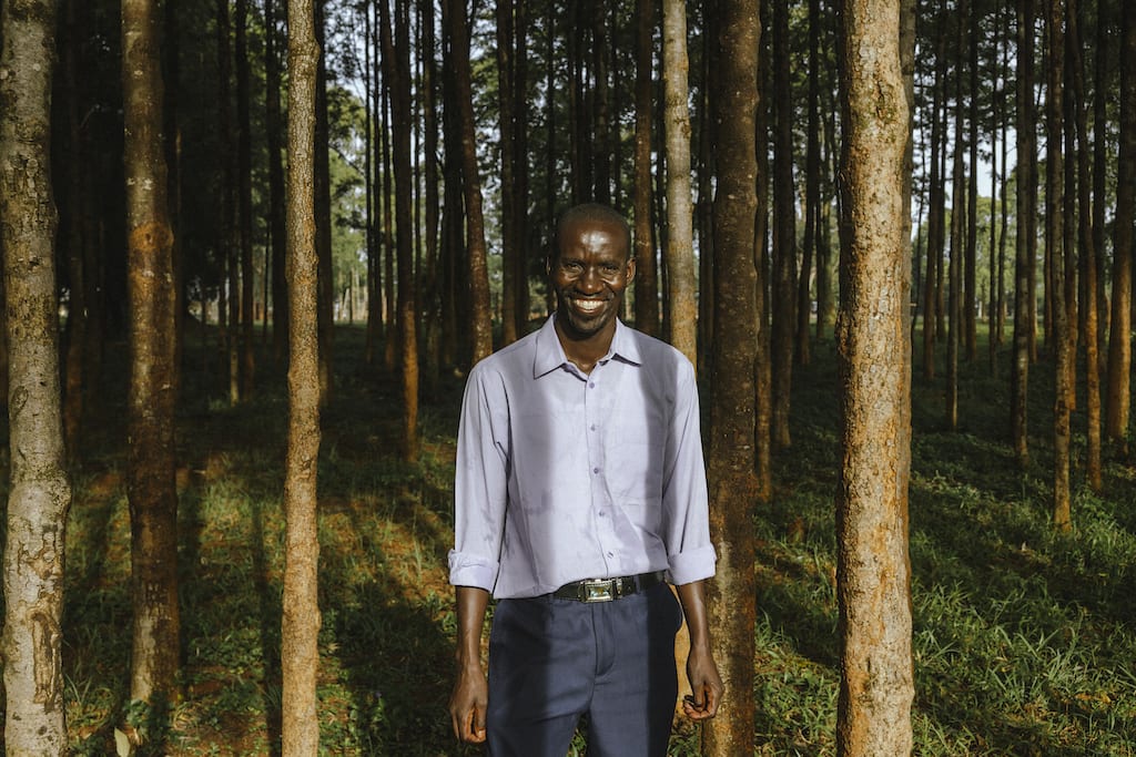Benson, the Project Director, wearing a light blue shirt and dark blue pants, is standing outside the center. There are trees all around him.