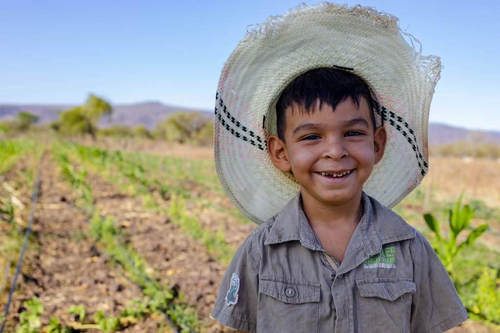Abner is wearing a tan shirt and a straw hat. He is standing outside in his family's garden.