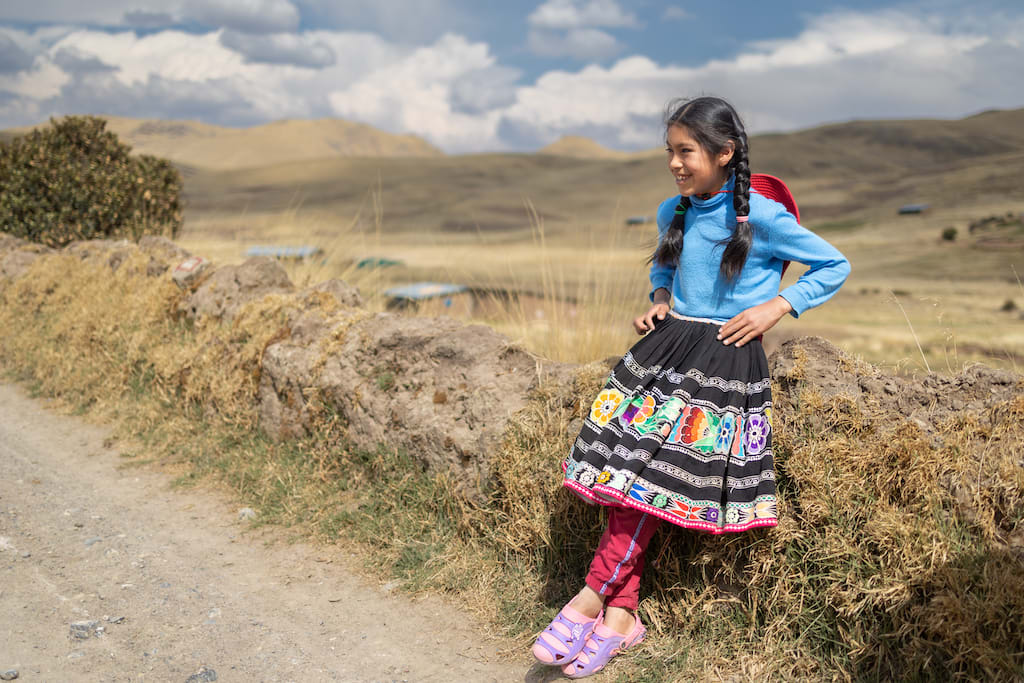 Young girl leans against a grass ledge smiling. She is wearing purple sandals, a colourful skirt, a blue shirt and braids in her hair.