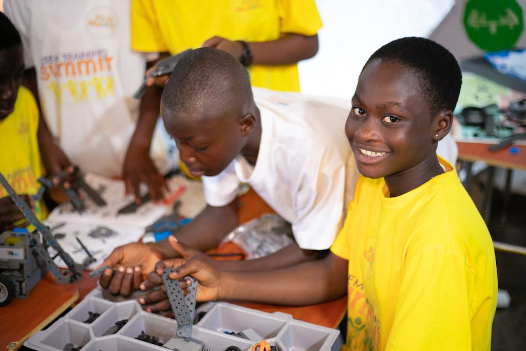 A girl in a bright yellow shirt gits with two small metal plates in her hand. Another girl in a white shirt leans over a container on the table.