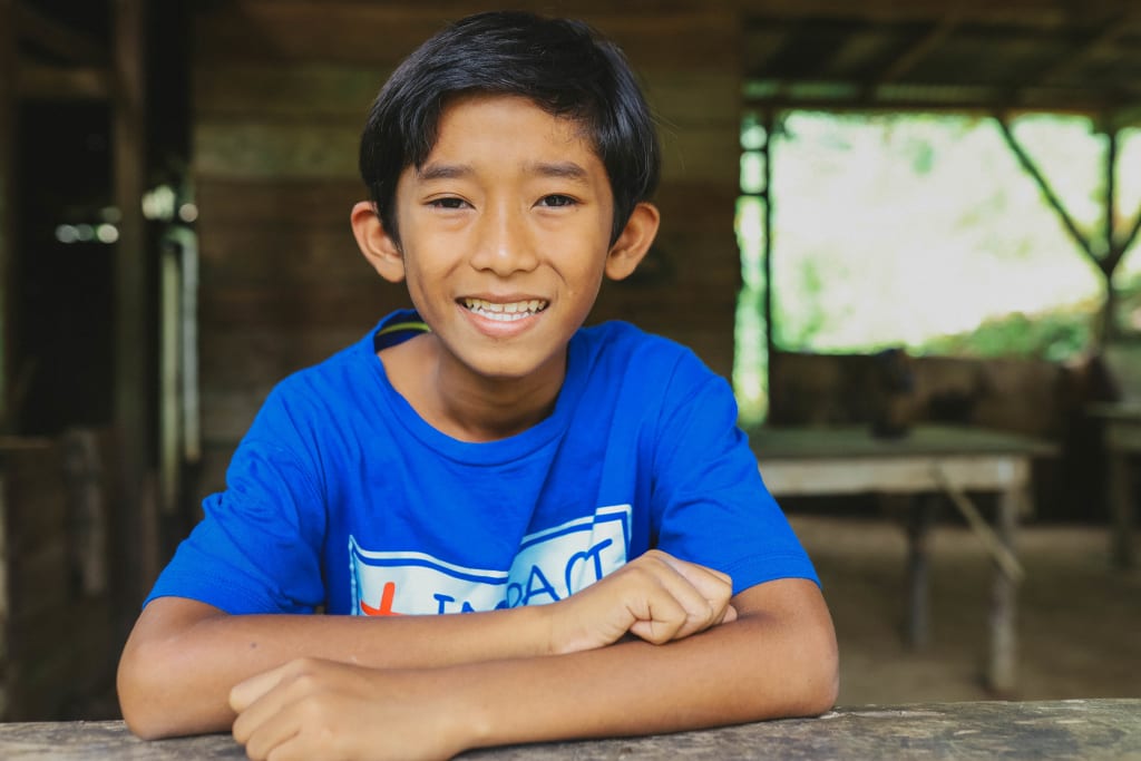 A boy in a bright blue t-shirt sits with his arms crossed on a table and smiles.