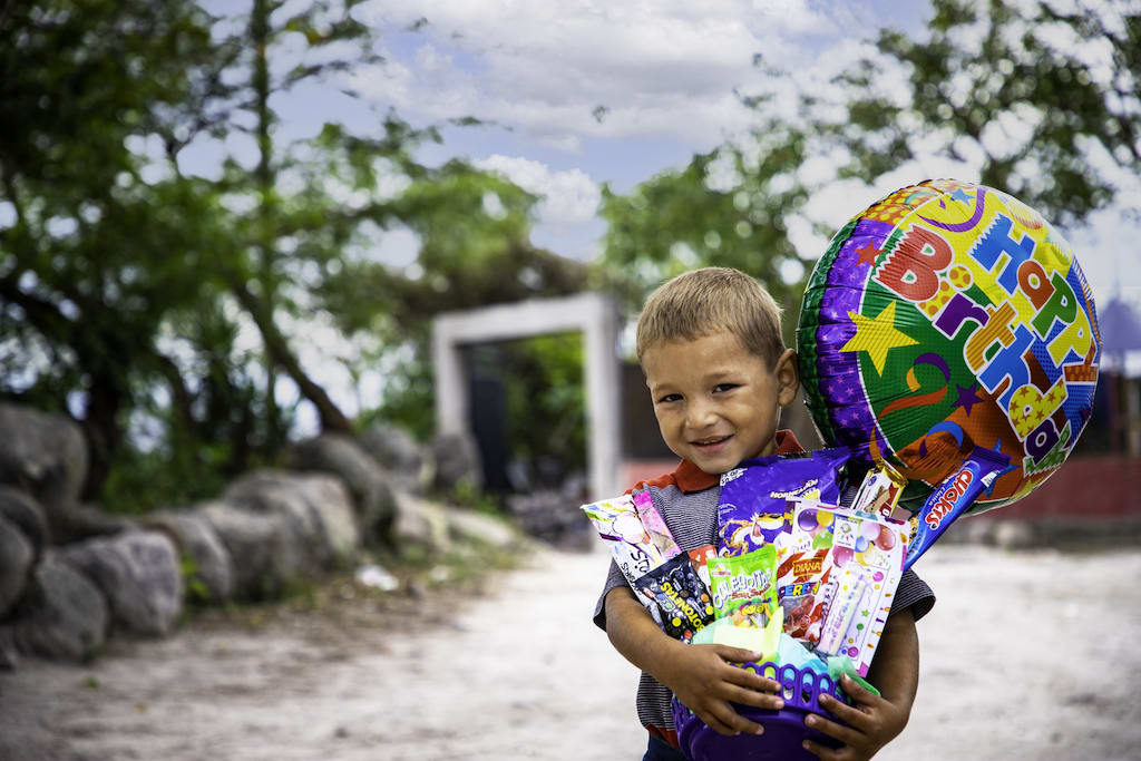 Mateo is wearing a gray shirt with a red collar. He is standing outside the Compassion centre holding a birthday gift basket he received.