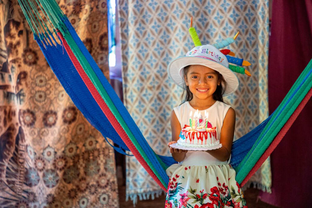 A girl in a colourful dress holds a cake with candles and wears a birthday hat