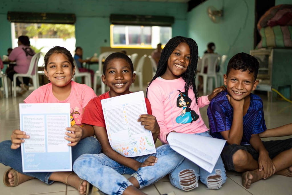 Four children sit on the ground smiling with letters in their hands.