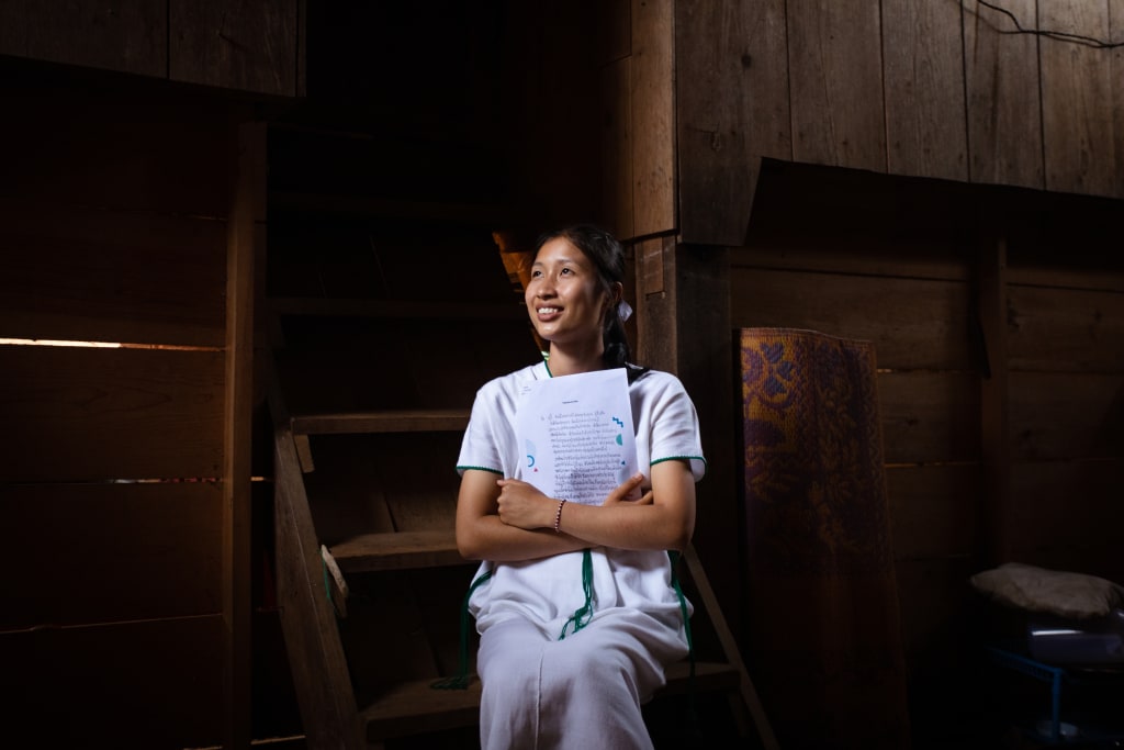 A teenage girl sits in a dark room wearing white and holding letters to her heart.
