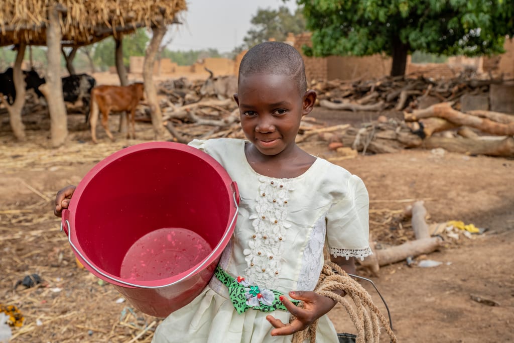 A girl in a white top stands with a big red bucket in her arms.
