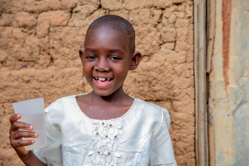 A girl in a white top stands with a white cup of water in her hand and smiles.