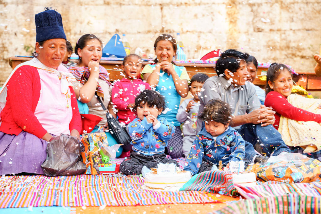 A group of people sit on the ground with confetti falling. There is a cake sitting in front of twin boys.