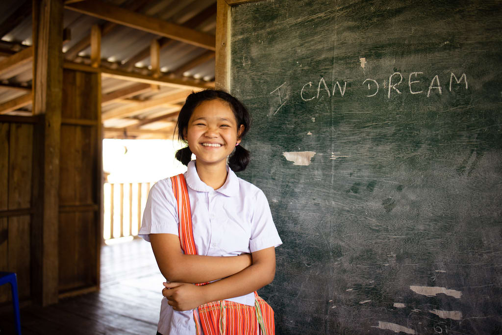 A girl is wearing a white shirt and an orange purse around her shoulder. She is standing in front of a chalkboard and smiling.