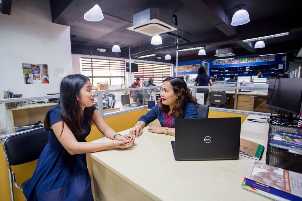 A girl in a blue dress sits across a desk from a woman in a blue blazer and they are talking