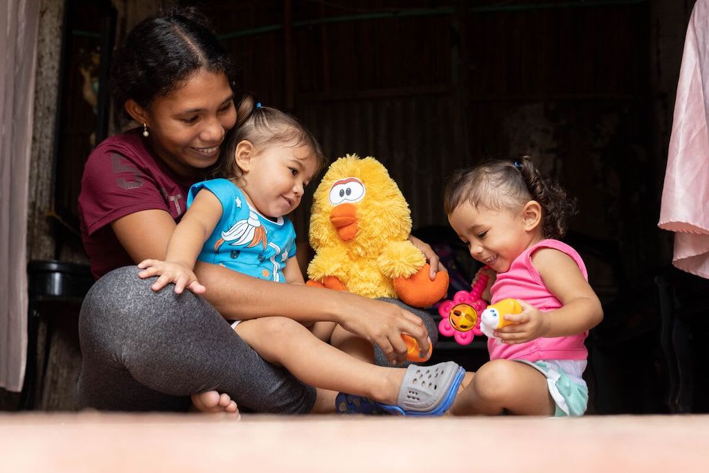 Salomé is wearing a pink shirt with blue pants. Ariana is wearing a blue shirt. They are sitting with their mother, Marta. She is wearing a maroon shirt and gray pants. They are holding a yellow stuffed animal.