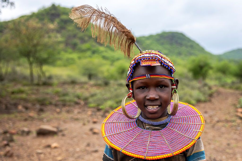 Salina is wearing a dress made with goatskin and a beaded head piece. She is standing outside her home. There are green hills and trees behind her.