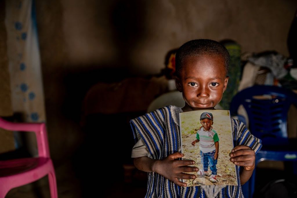 Achille is wearing a blue tunic and is holding a photo of his younger self.