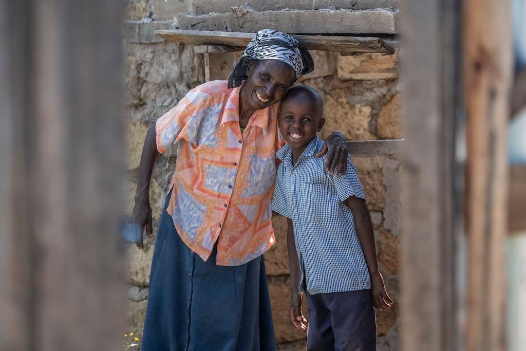 Nkatha and Brian stand outside their home, smiling. Nkatha's arm is around Brian's shoulders.