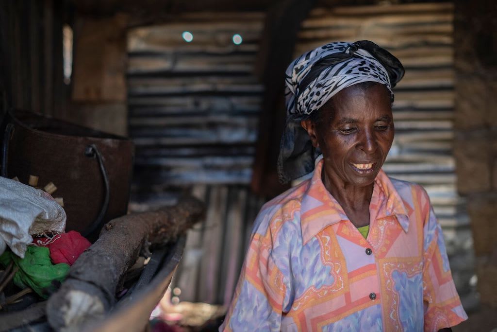 Nkatha stands in her home, wearing a black and white headscarf and a patterned peach shirt.
