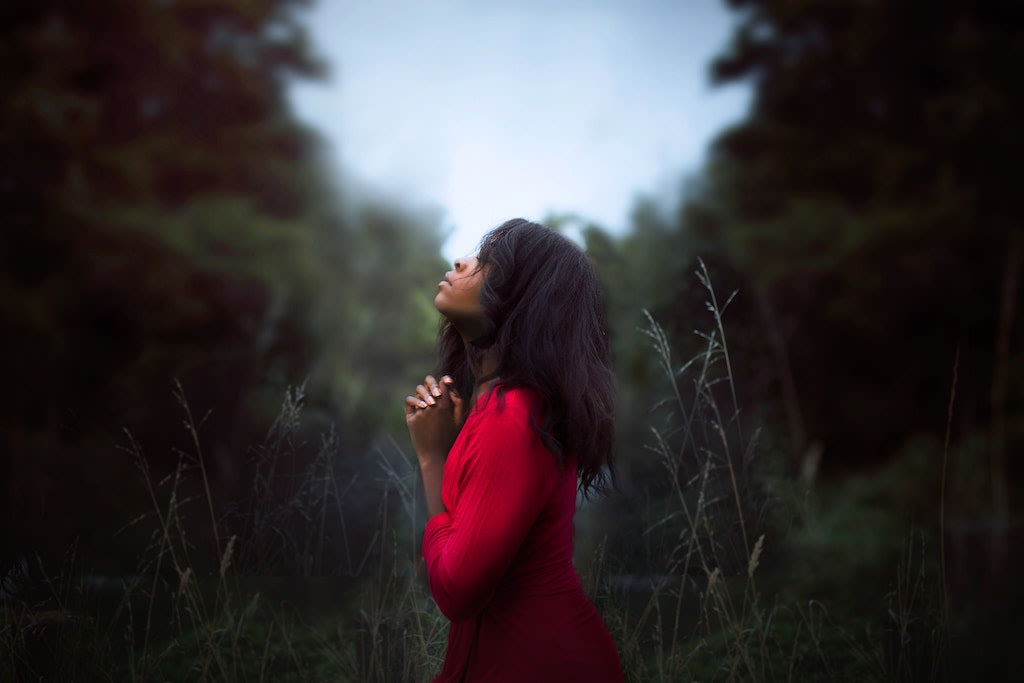 A woman standing in a field with her hands folded and looking upwards.