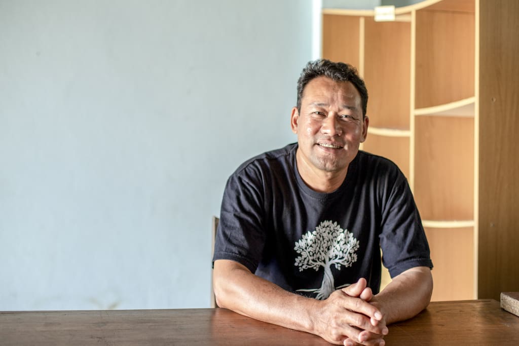 Somsak sits at a desk in a black t-shirt, looking at the camera and smiling warmly.