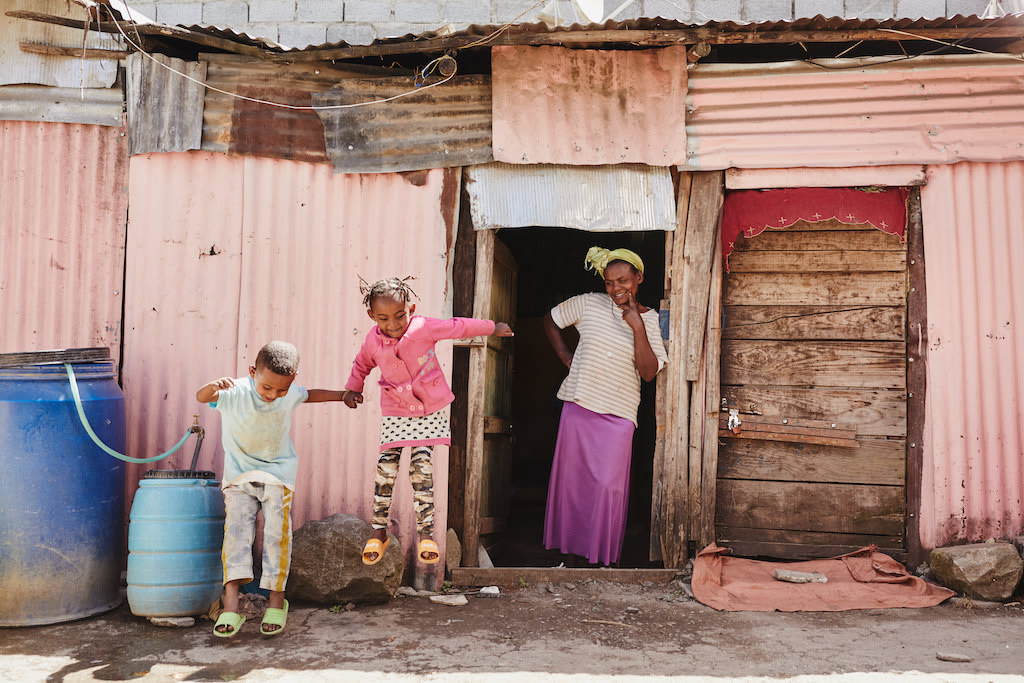 A mother stands in the door of her home, watching her two young children jump.