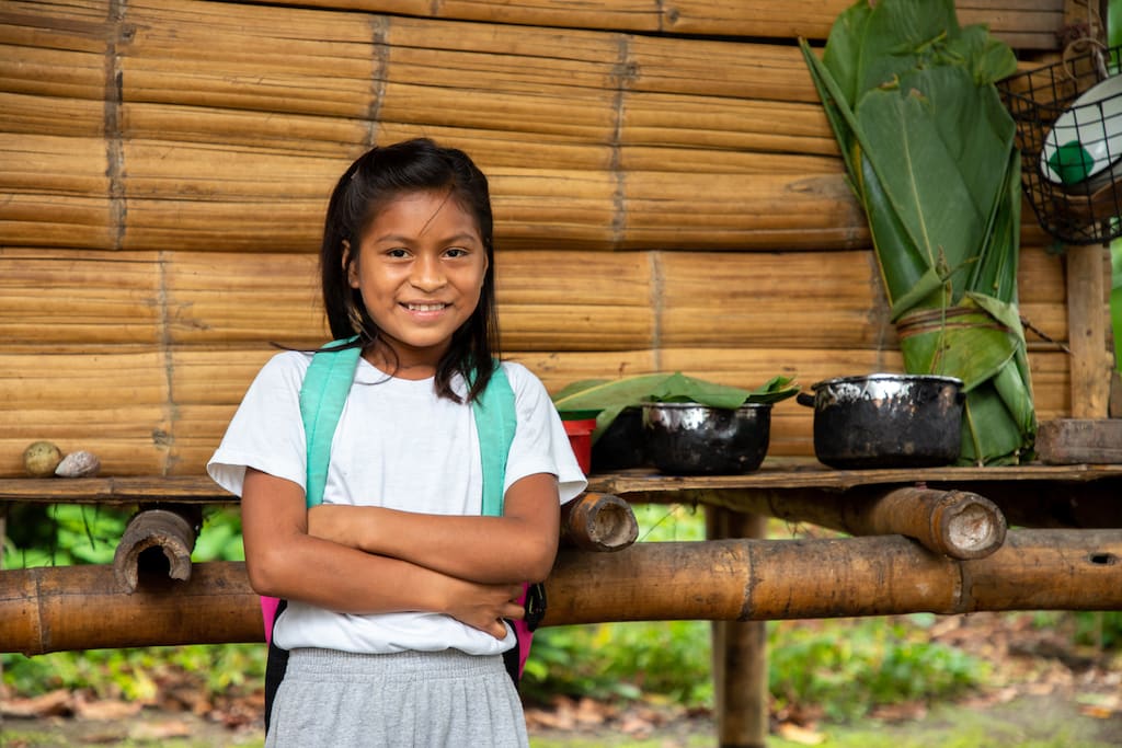 A girl poses with her arms folded, she is wearing a teal backpack and white t-shirt.
