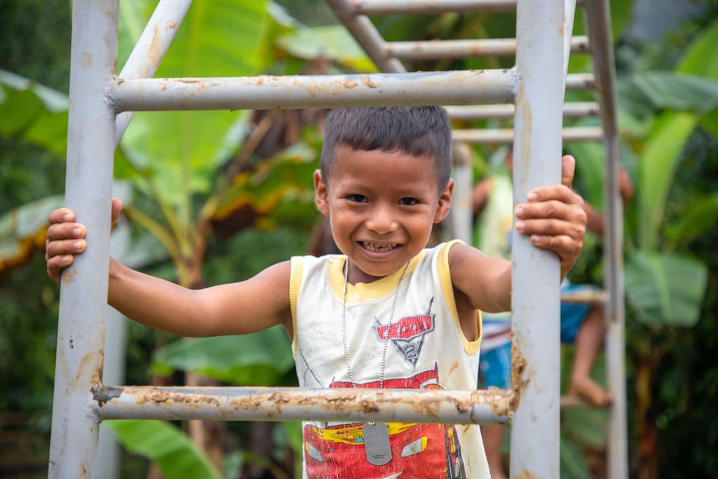 Through a playground ladder, a young boy wearing a white and orange tank top smiles at the camera.