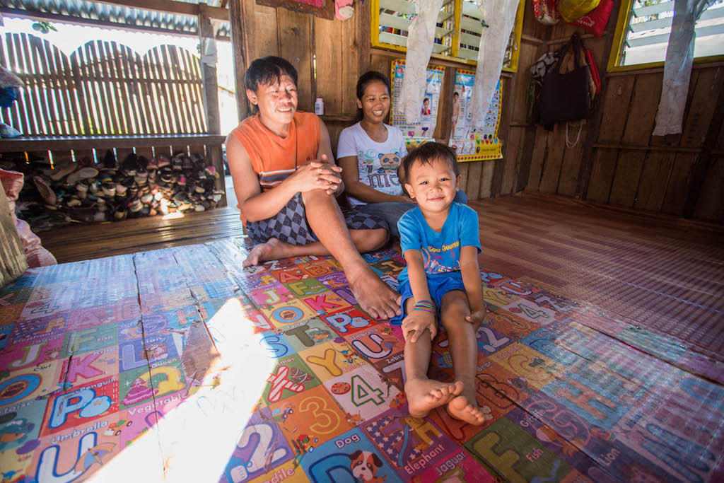 Jandell and his parents sit on the floor of their home. Jandell looks at the camera while his parents sit behind him looking at him.