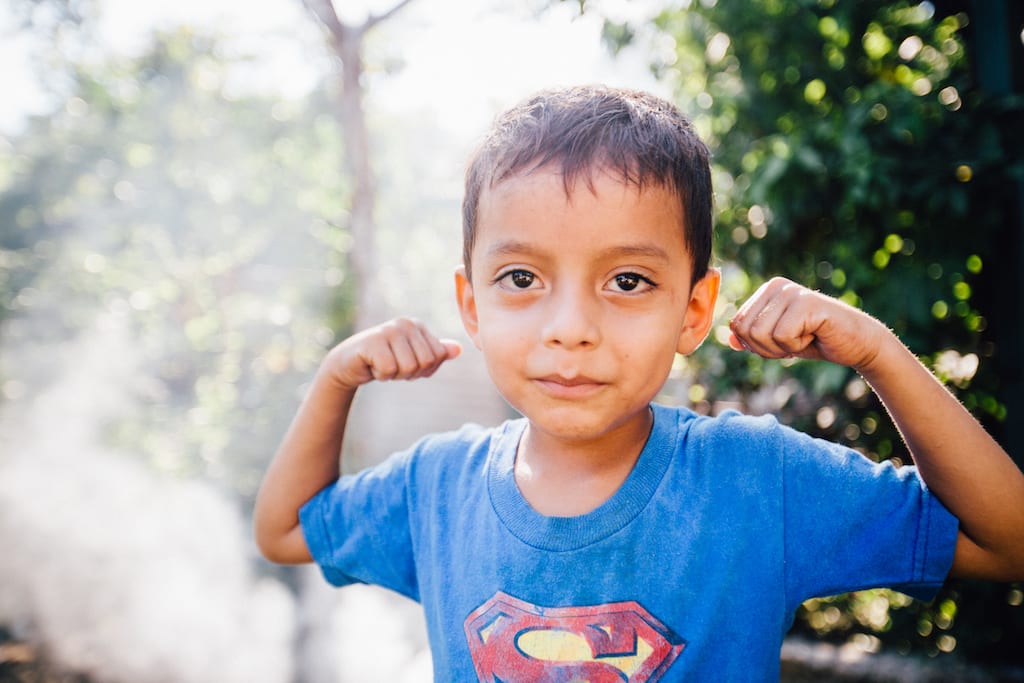 A young boy in a blue t-shirt stands flexing his arms.