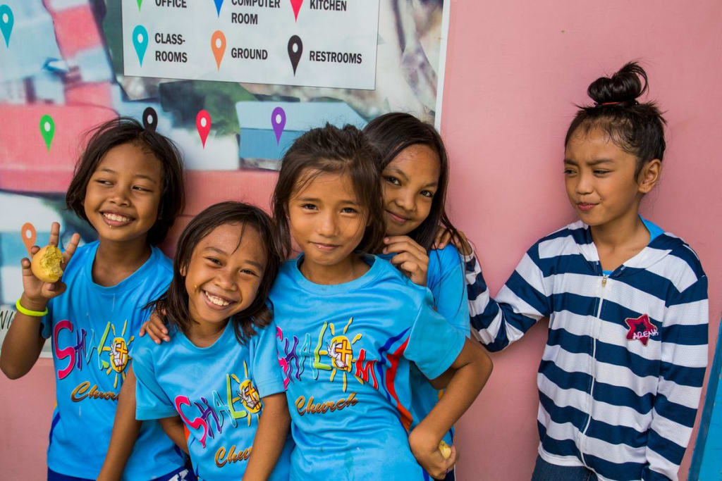 A group of girls smile together wearing matching blue shirts. There is a pink wall behind them.