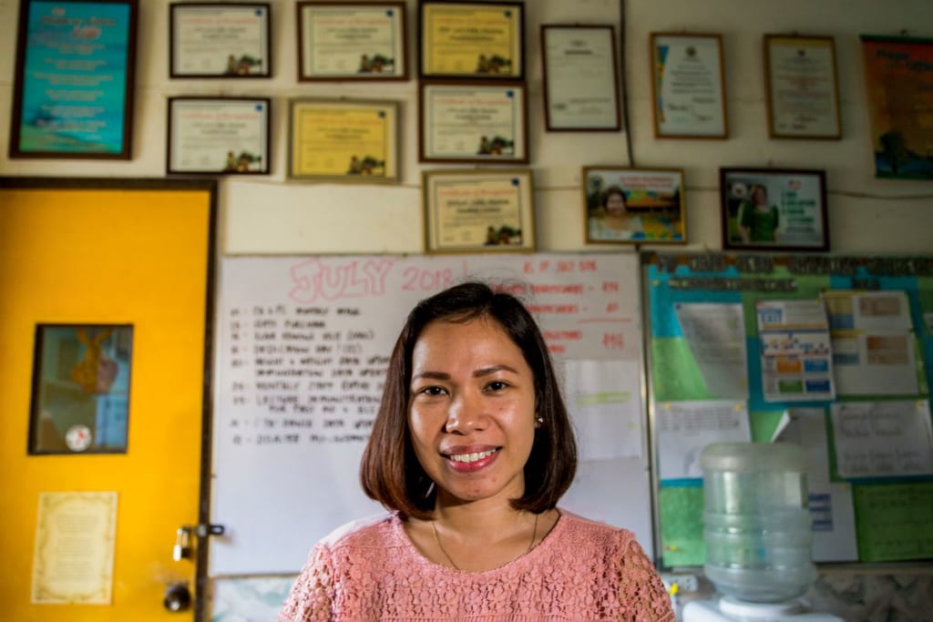 Diane smiles at the camera in a classroom, with awards and pictures behind her on the wall.